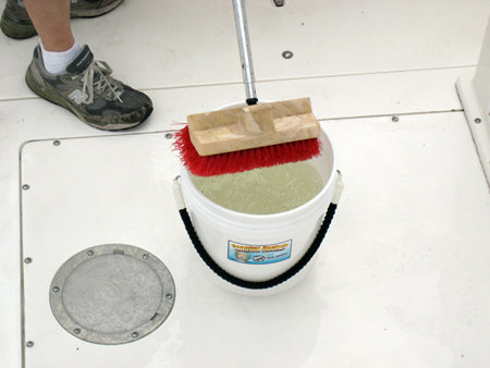 Bucket with cleaning brush and sponge on a white floor, with a person's feet in the background.